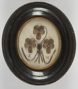 Composición formada con mechones de cabello sobre fondo de cartón blanco, representando un ramo con tres flores de cinco pétalos y lazo central.